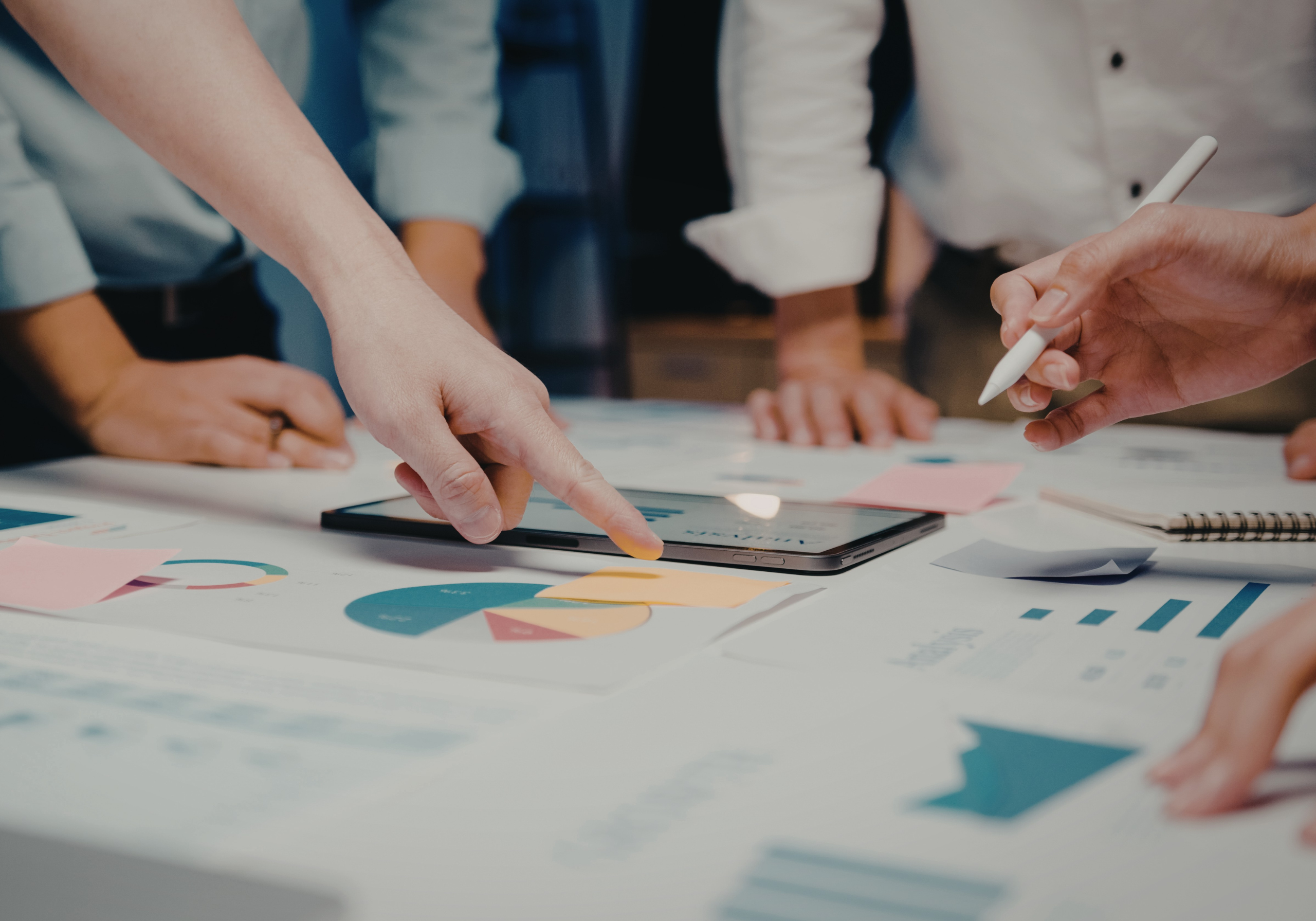 Un primo piano sfocato di più paia di mani che esaminano attentamente vari documenti stampati, report e mappe sparsi su un tavolo da ufficio. Una mano tiene una penna e un'altra indica una sezione di un grafico